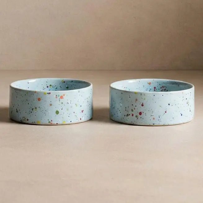 Twee Soft Blue Splatter voerbakken van steengoed met veelkleurige spikkels tegen een beige achtergrond